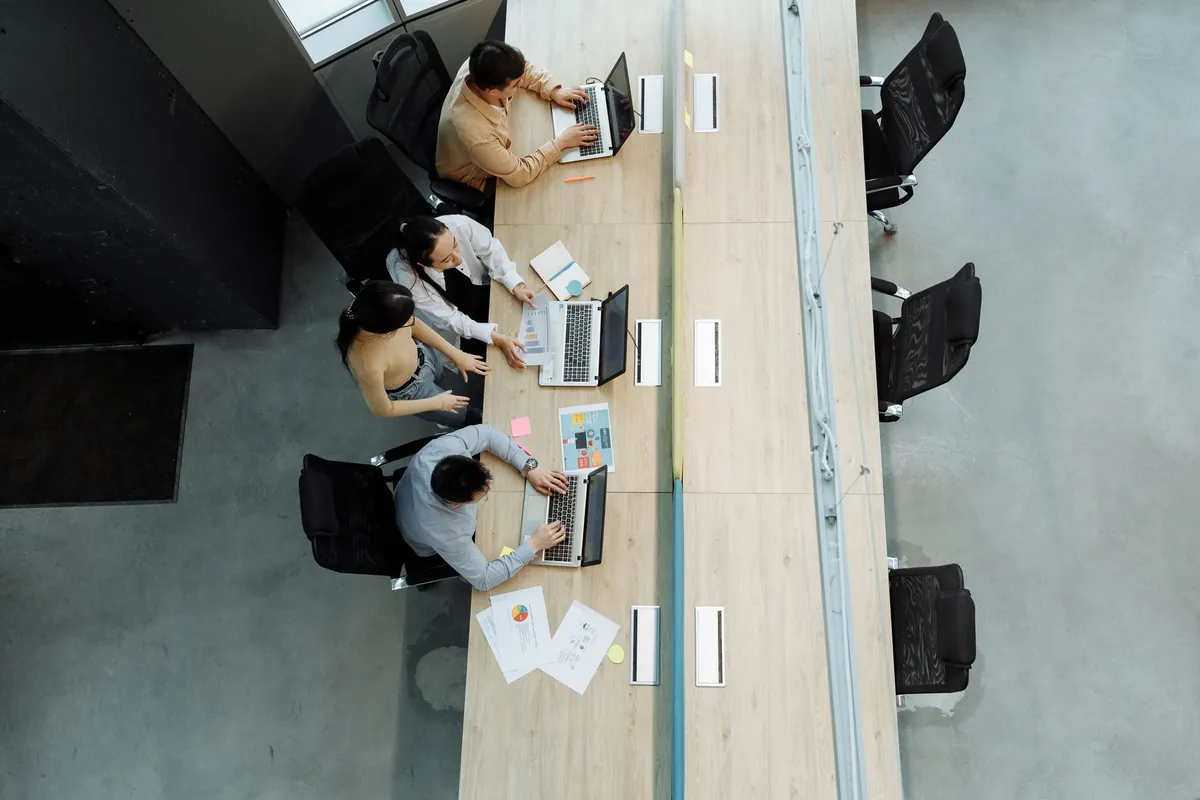 An open office seen from above, a team working side by side at long shared desks with laptops and papers