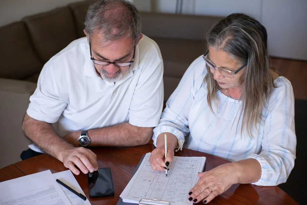 An older couple at a wooden table going through printed papers together, one of them holding a phone
