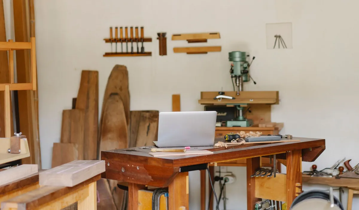 A small woodworking shop with a laptop open on the workbench, timber stacked against the wall